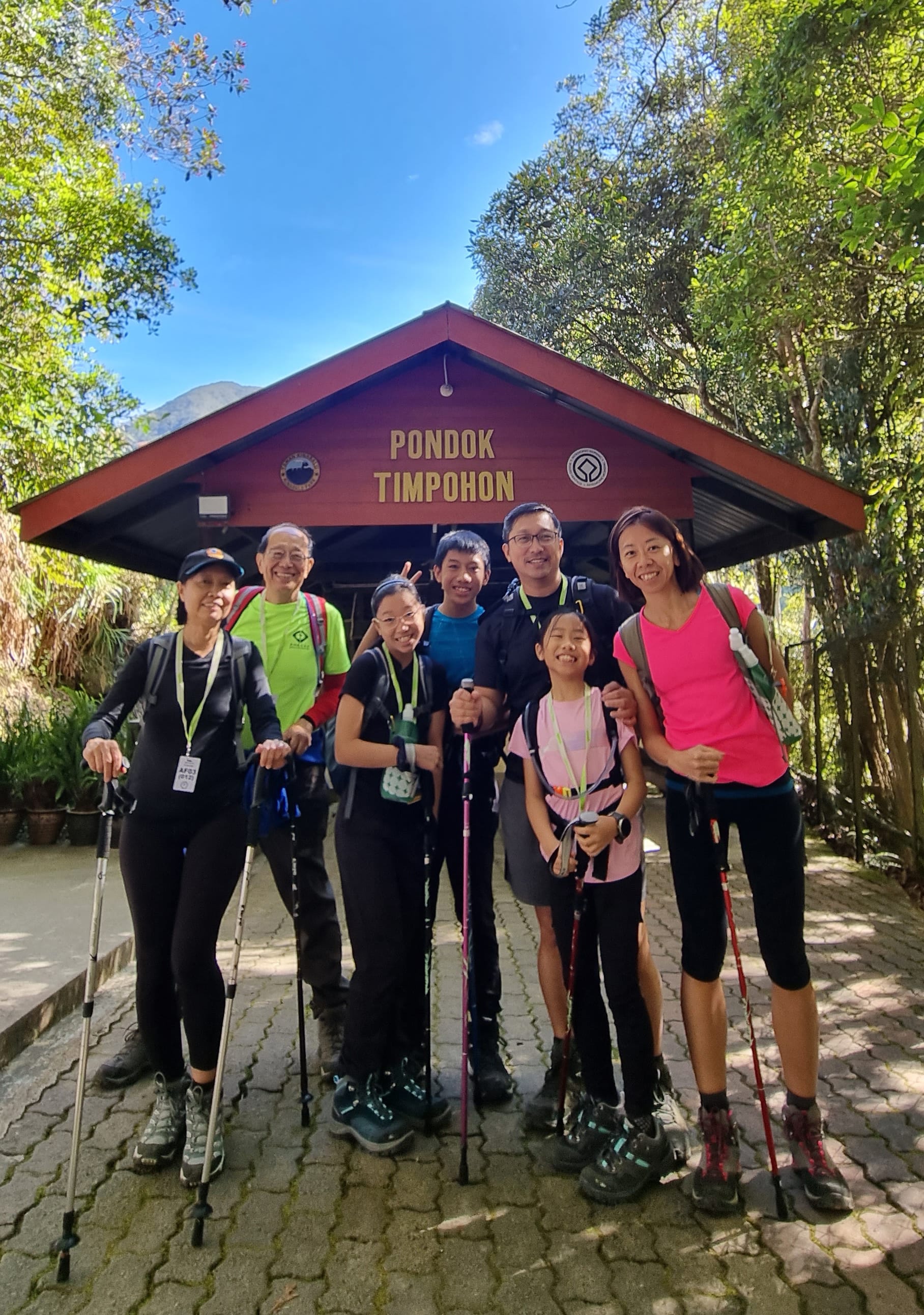 Group of hikers standing outside Pondok Timpohon hut with trees and bright blue sky in the background.